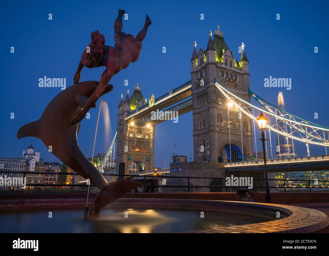 The first light of the morning blue hour begins to illuminate the iconic Girl With A Dolphin Fountain, Tower Bridge and The Shard in Central London. Stock Photo