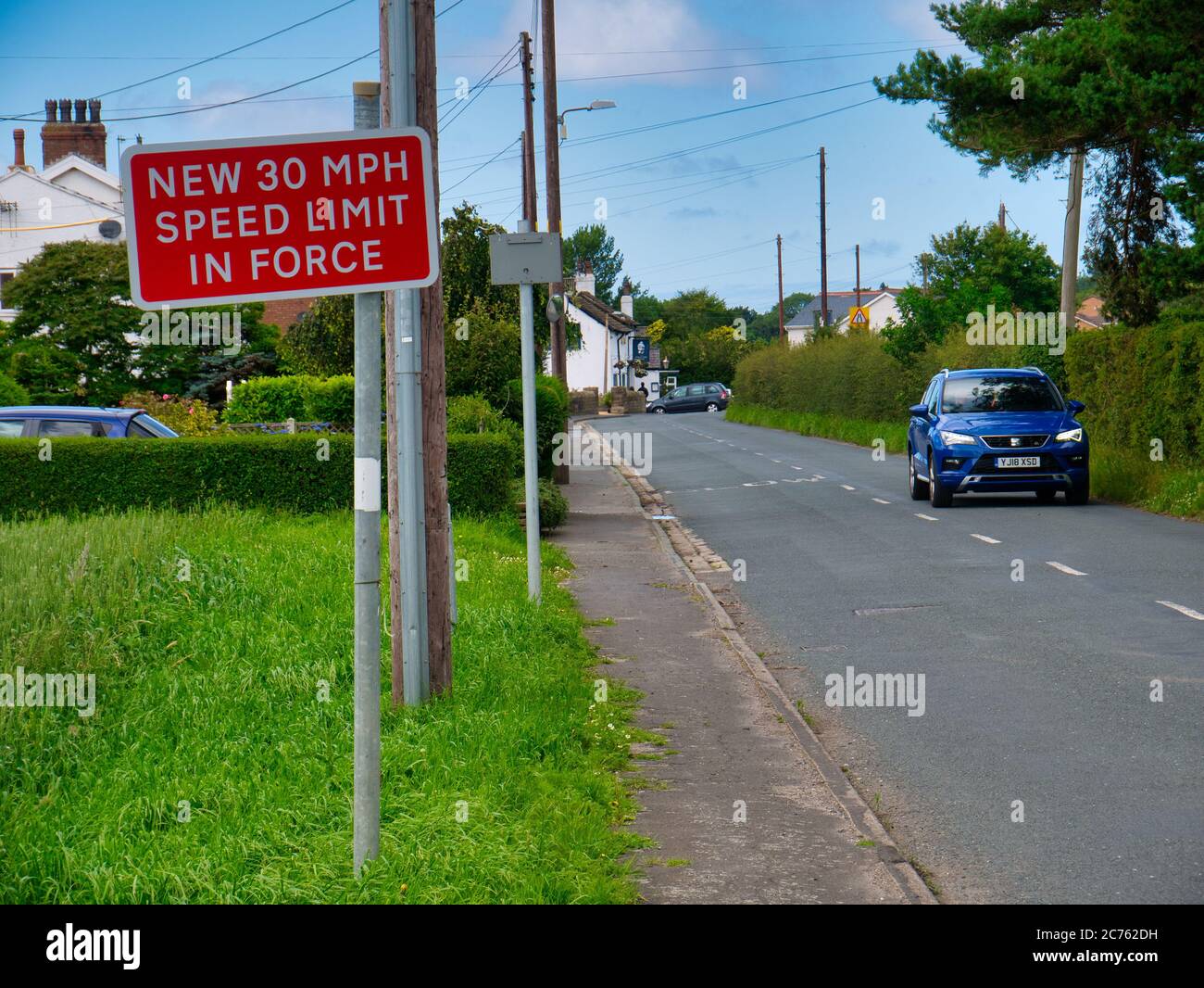 A red sign warns motorists of a new 30mph speed limit in force in the rural village of Halsall in Lancashire, UK. A blue car passes on the right. Stock Photo