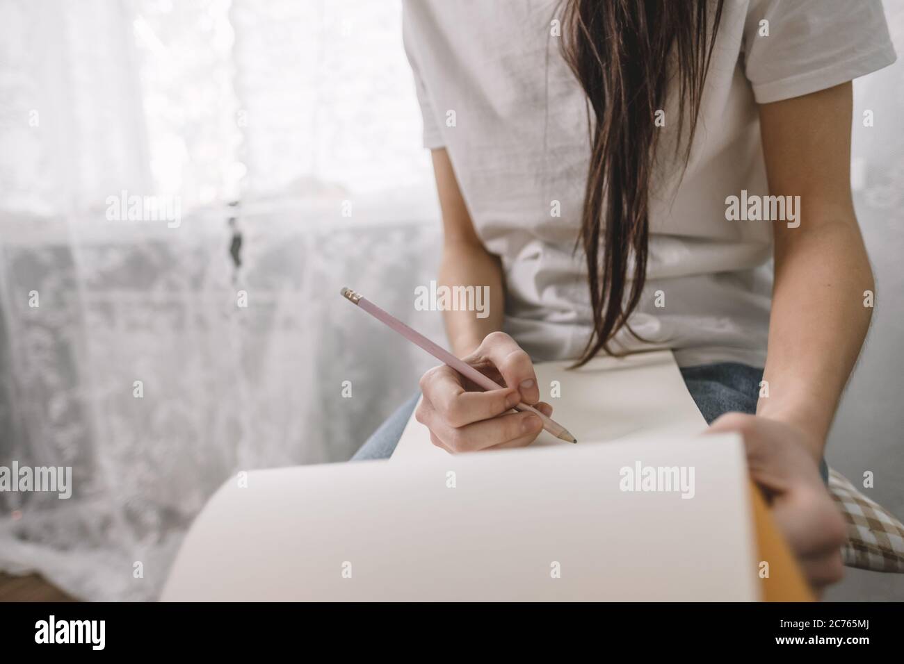 Woman artist draws in a sketchbook at home near the window, sitting on a chair Stock Photo