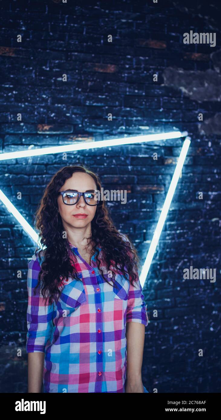 Young woman with glasses in colored plaid shirt posing on background shabby brick wall with glowing lamps. Portrait of charming female against Stock Photo