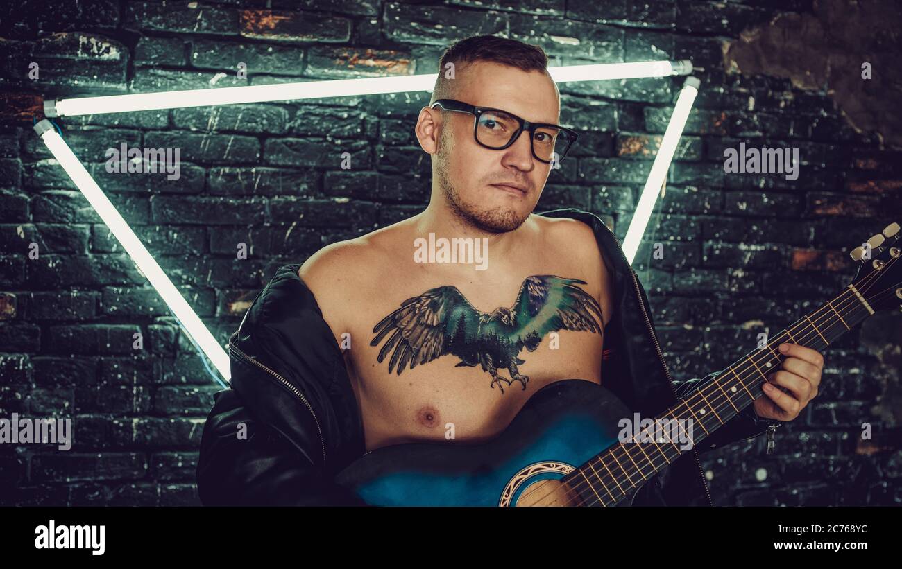 Tattooed man playing guitar near illuminated wall. Stylish guy with bird tattoo on chest looking away and playing guitar while standing against shabby Stock Photo