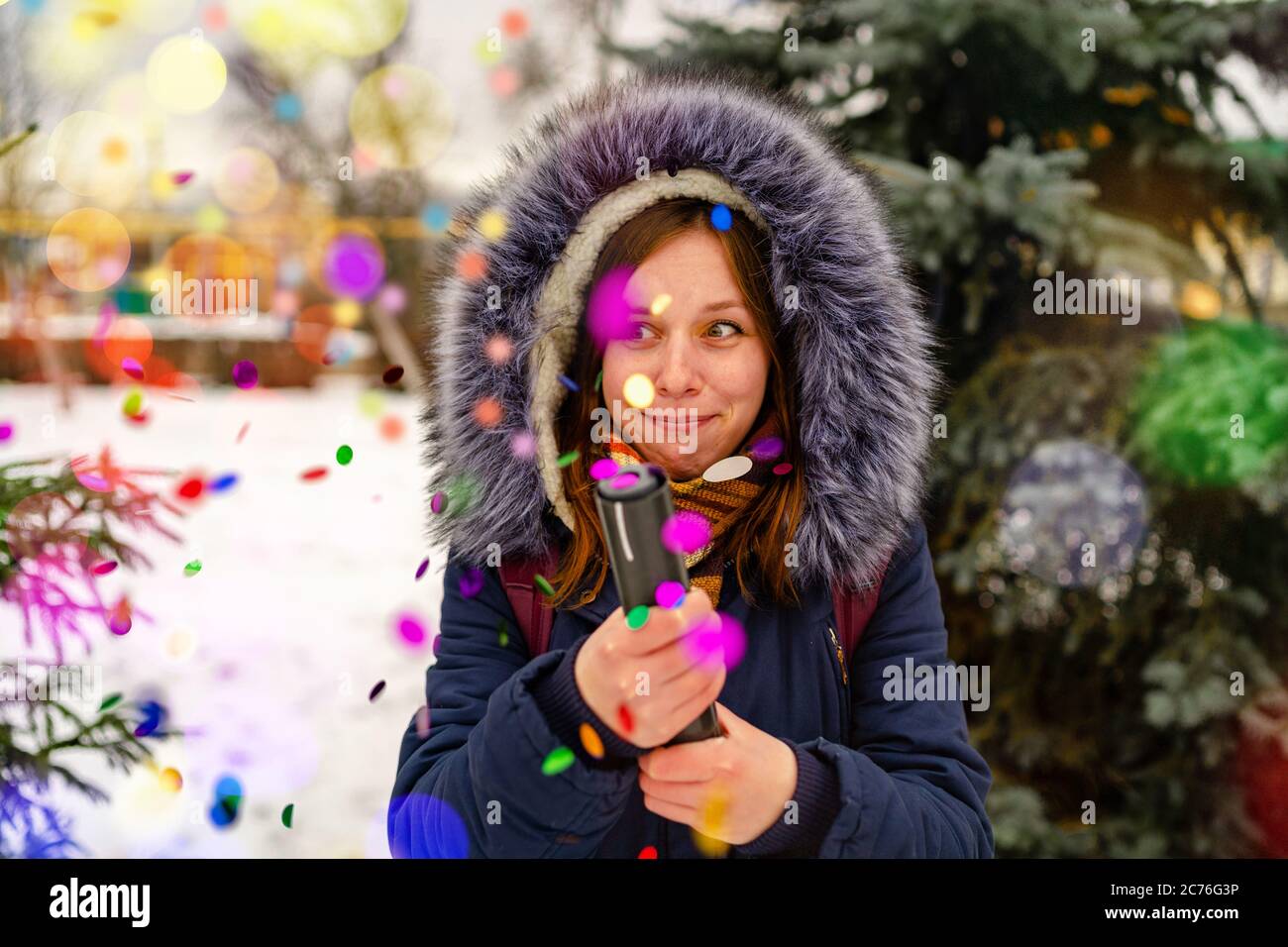 Emotional woman pops confetti petard and opens her mouth with delight. Adult female in hooded jacket admires and pops confetti petard into camera Stock Photo