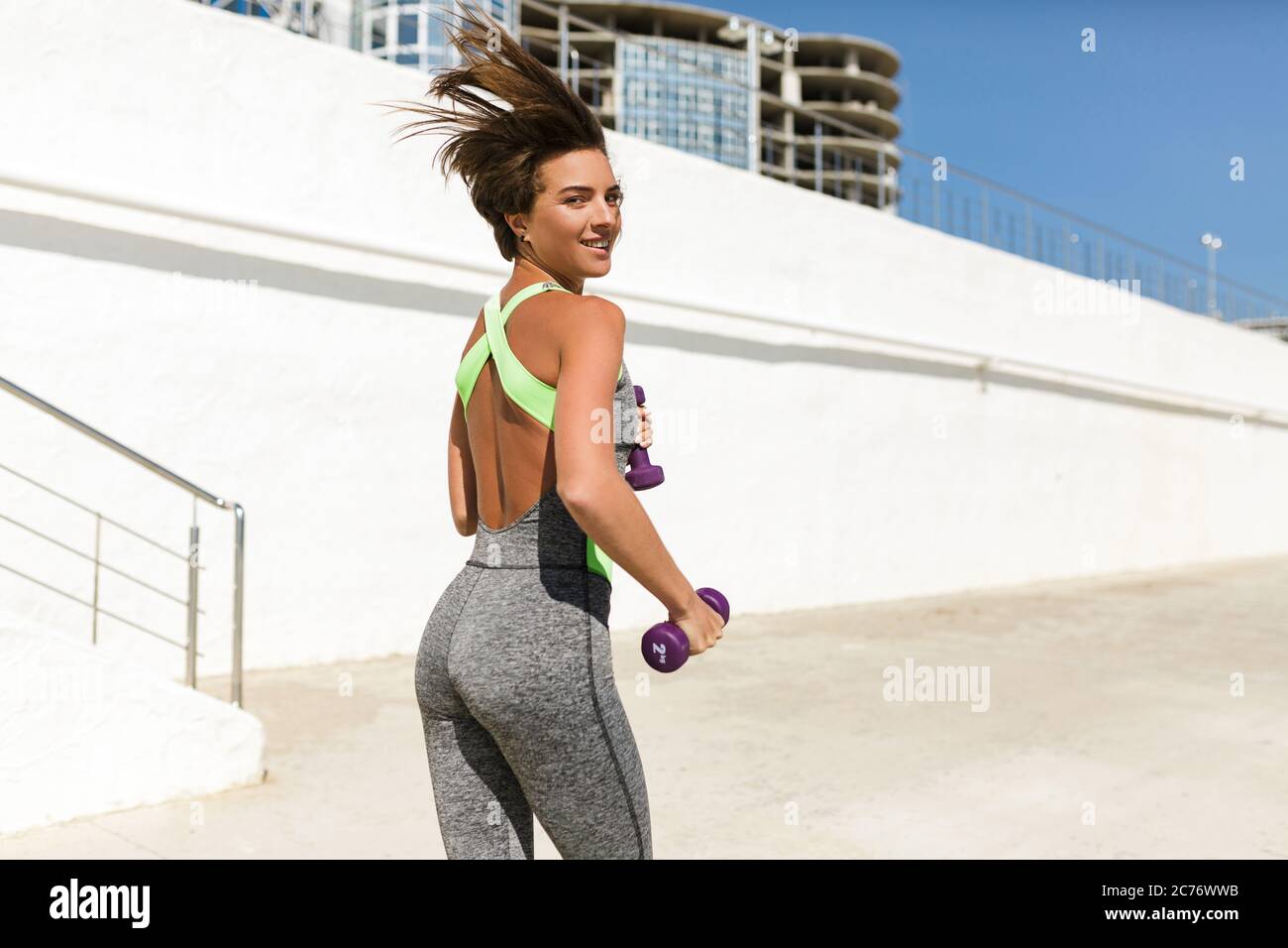 Beautiful woman in modern sport suit holding dumbbells and jogging joyfully looking in camera Stock Photo