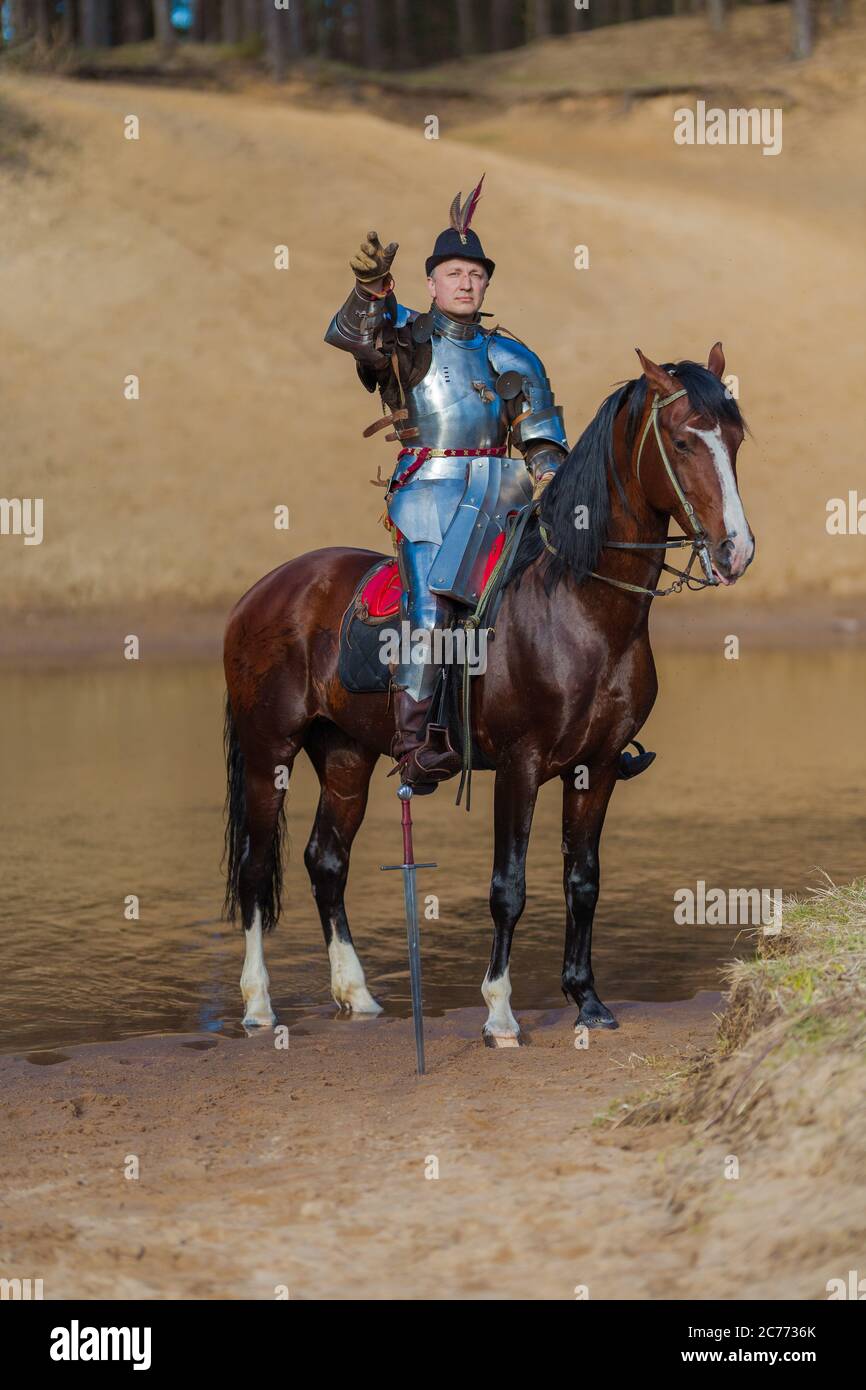 Adult man in ancient knight armor with a sword rides a horse on a river ...