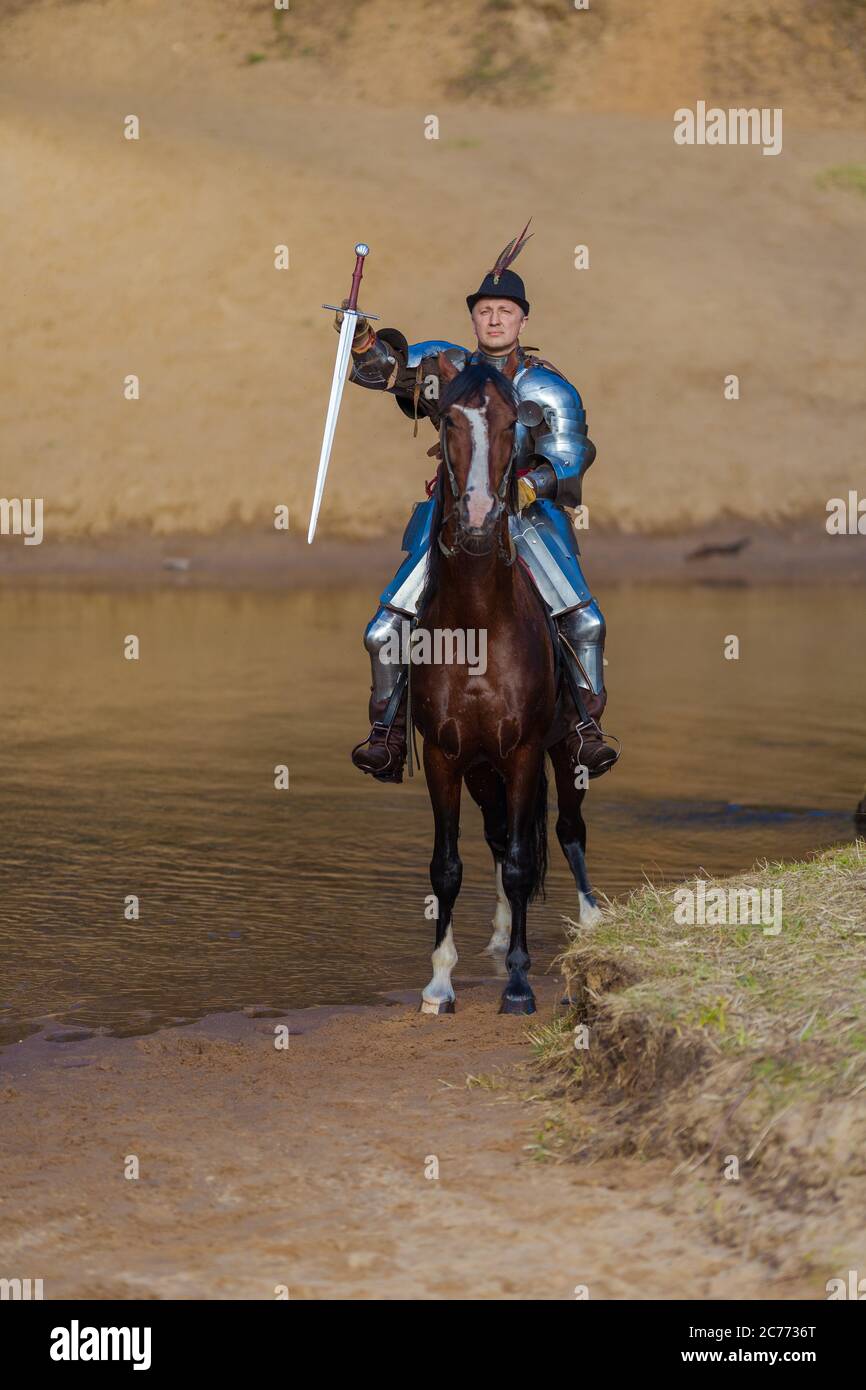 Adult man in ancient knight armor with a sword rides a horse on a river ...
