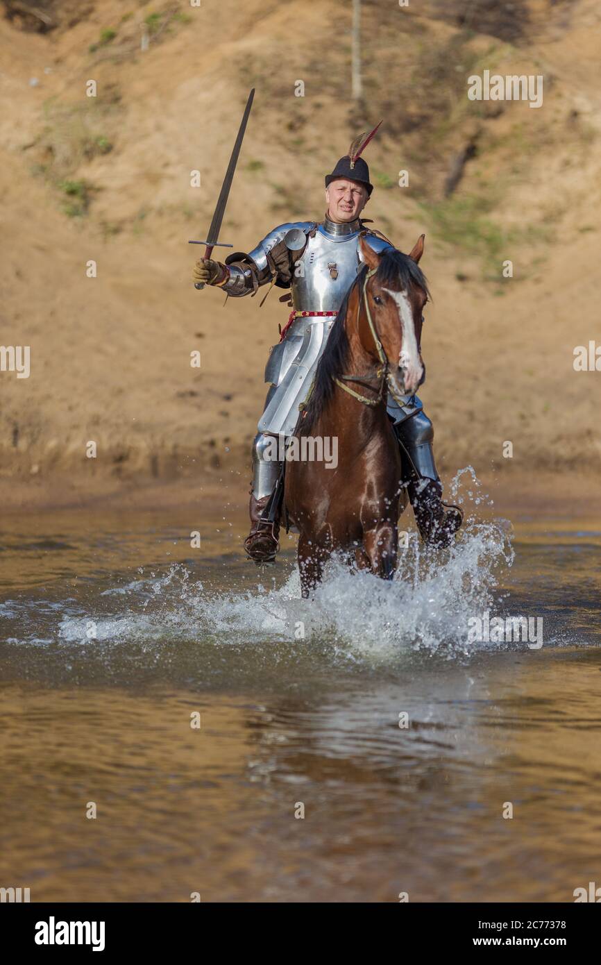 Adult man in ancient knight armor with a sword rides a horse on a river ...