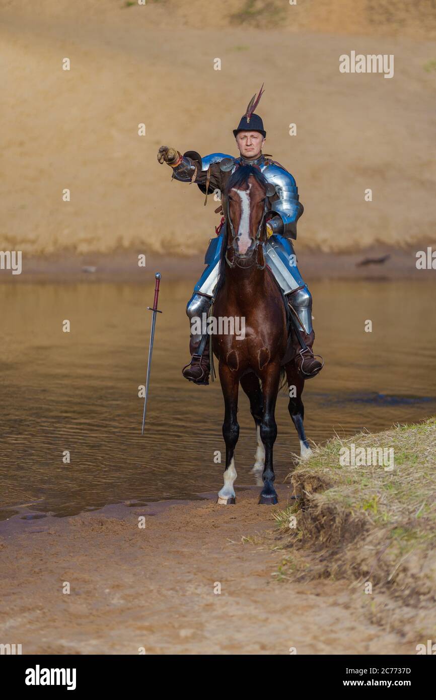 Adult man in ancient knight armor with a sword rides a horse on a river ...