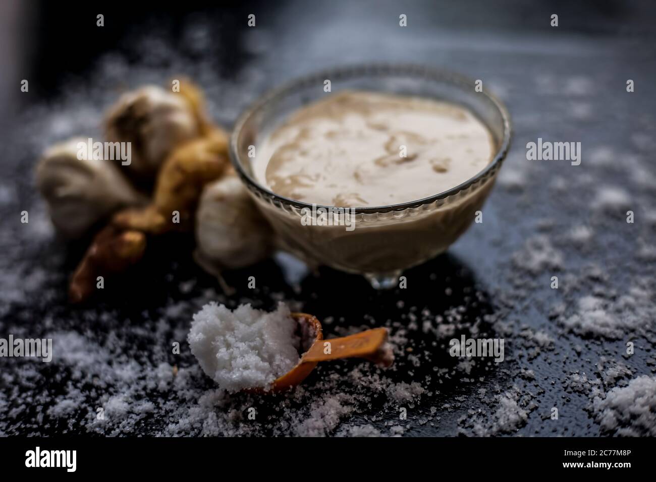 Shot of freshly ground garlic and ginger paste in a glass bowl on a black surface along with raw ginger, and some garlic with salt on sprinkle on the Stock Photo