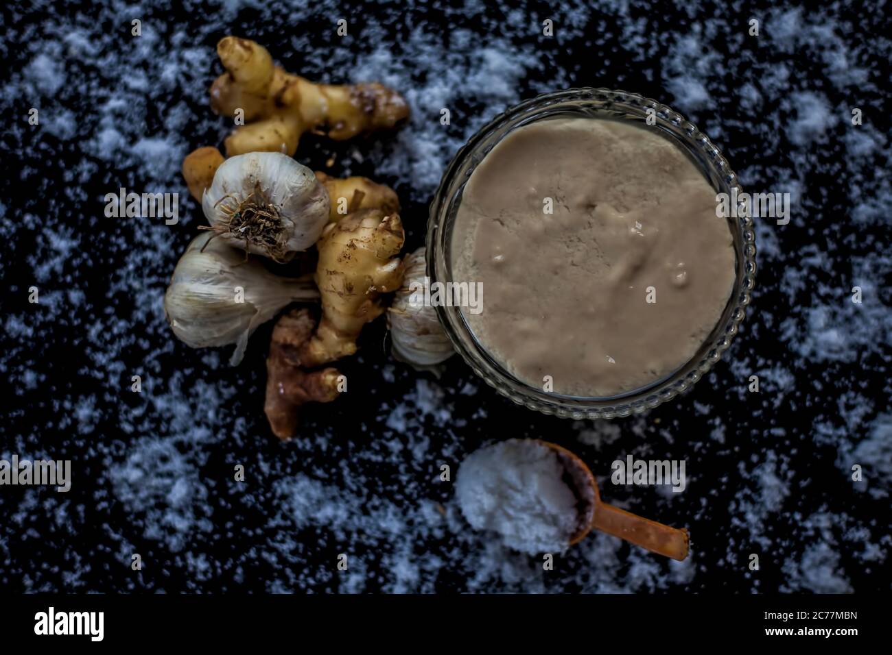 Shot of freshly ground garlic and ginger paste in a glass bowl on a black surface along with raw ginger, and some garlic with salt on sprinkle on the Stock Photo