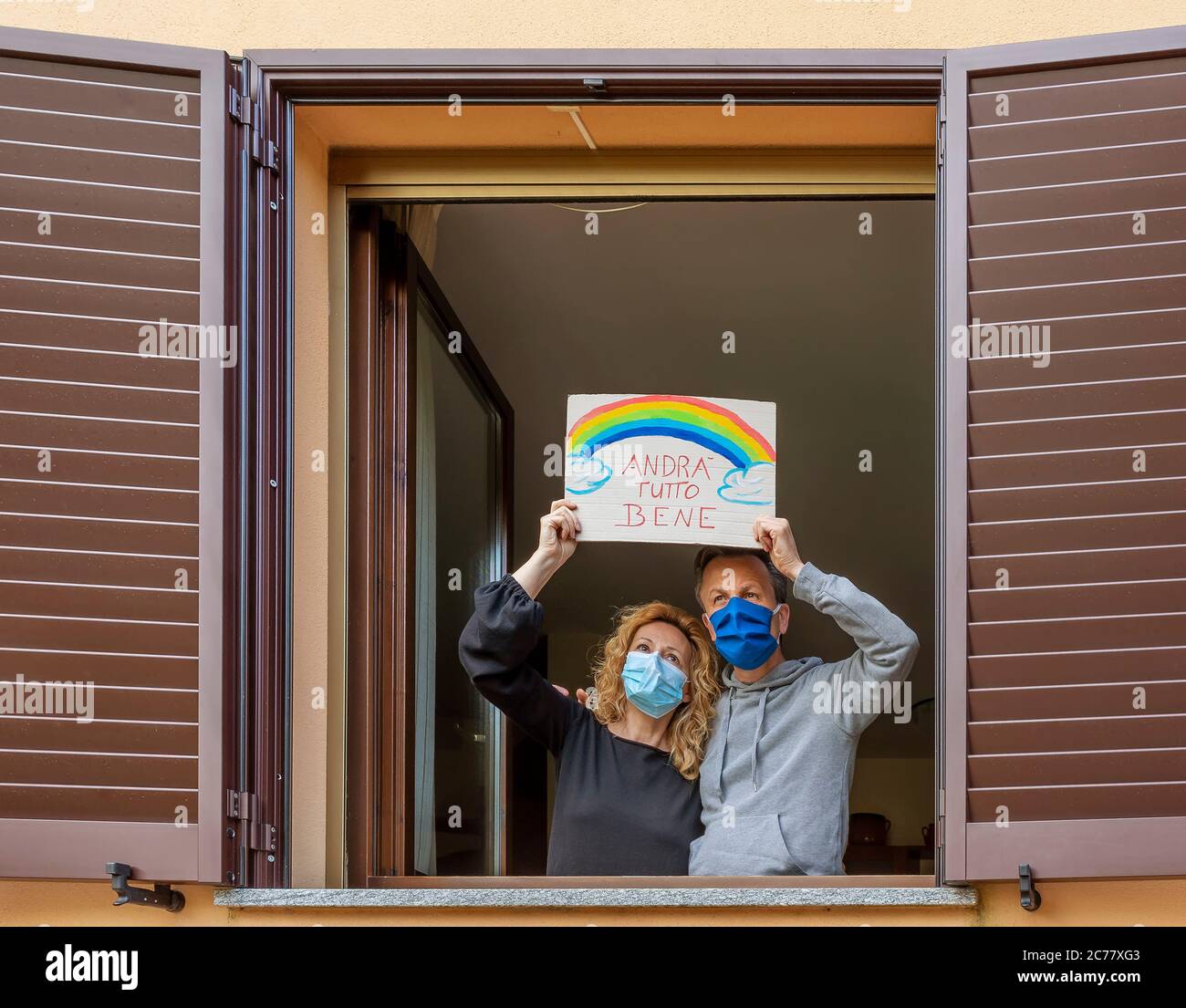 A middle-aged couple looks out the window with protective masks and exposes the sign 'Andra' tutto bene' which in italian means that everything will Stock Photo
