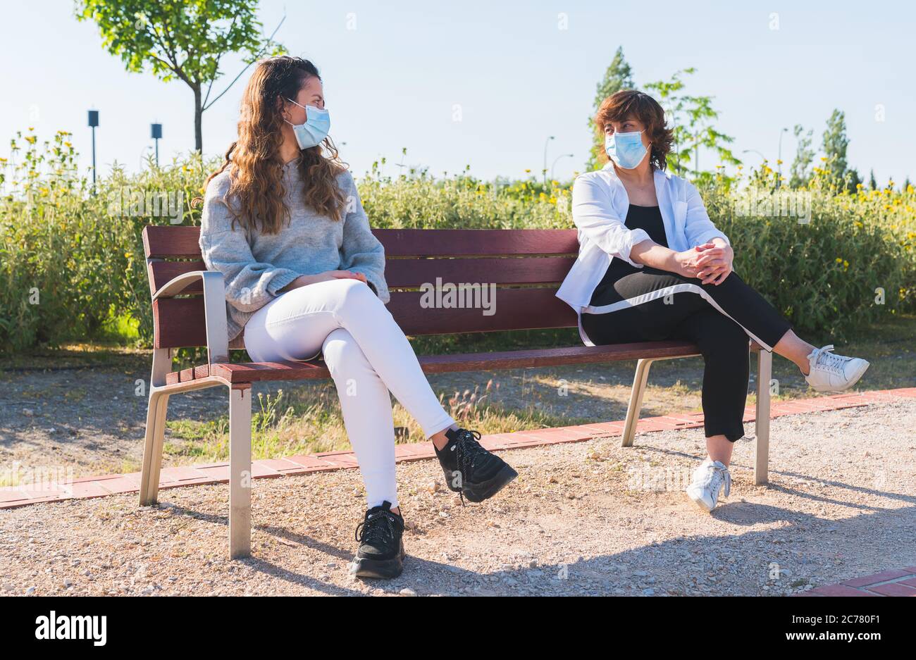 Social distance. Mother and daughter in social distance sitting on a park bench. Pandemic. Coronavirus. Maintaining safe distance sitting on a park be Stock Photo