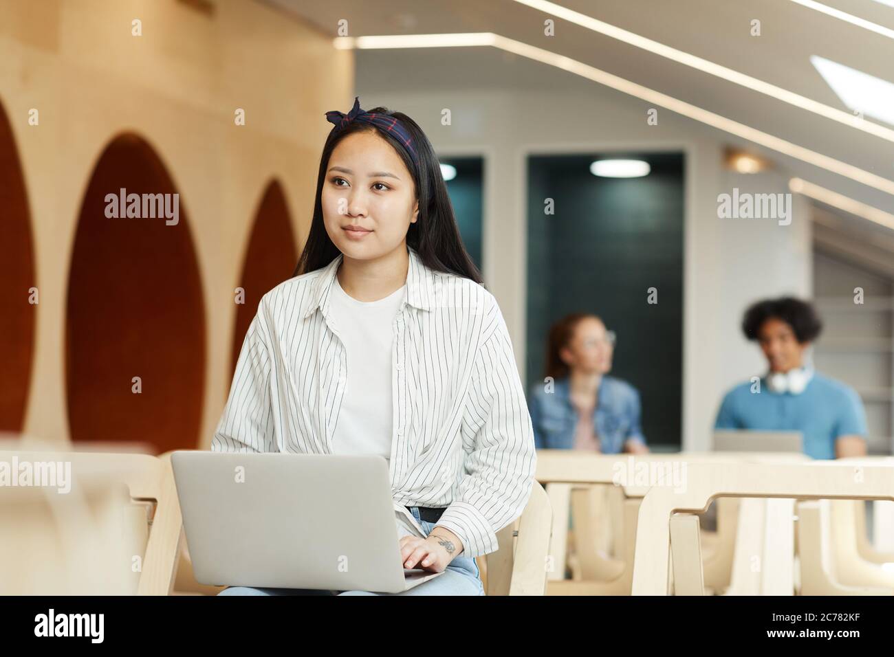 Content Asian student in headscarf sitting on chair in auditorium and using laptop at lecture Stock Photo