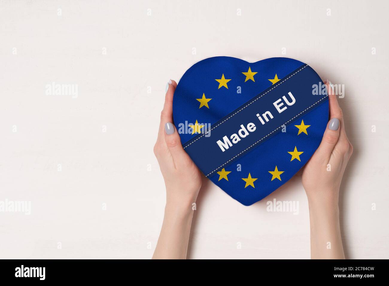 Inscription Made in EU, the flag of EU. Female hands holding a heart shaped box. White background. Place for text Stock Photo