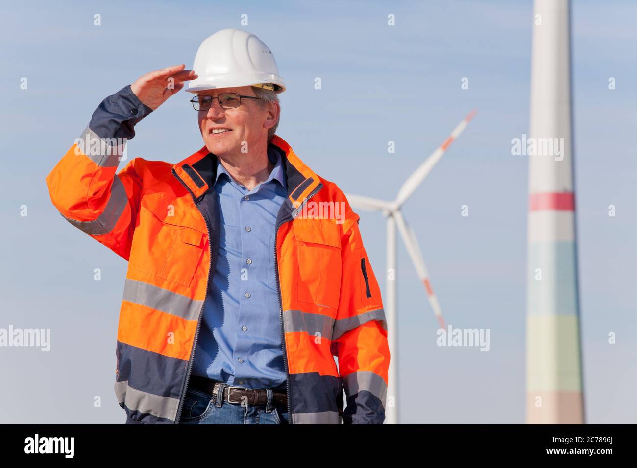 Engineer with hard hat and protective clothing in front of a windfarm looking for a bright future for green energy - focus on the person Stock Photo