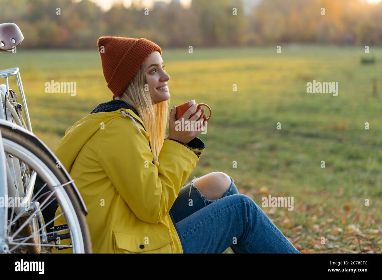 Beautiful smiling woman sitting on grass in park, holding cup of coffee. Travel, autumn concept Stock Photo
