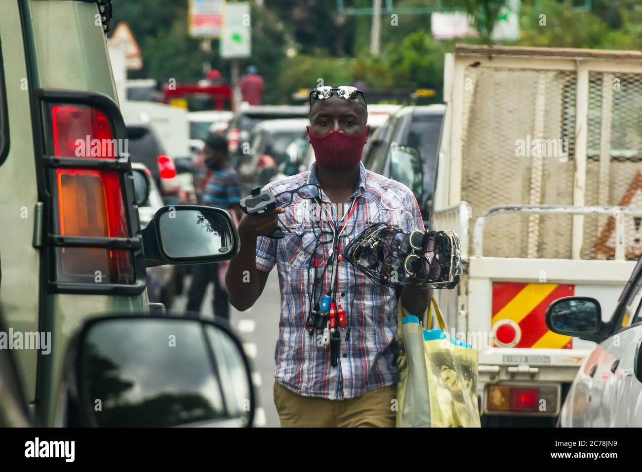A street hawker wearing a face mask walks past vehicles stuck in traffic selling sunglasses and other goods, Nairobi, Kenya Stock Photo