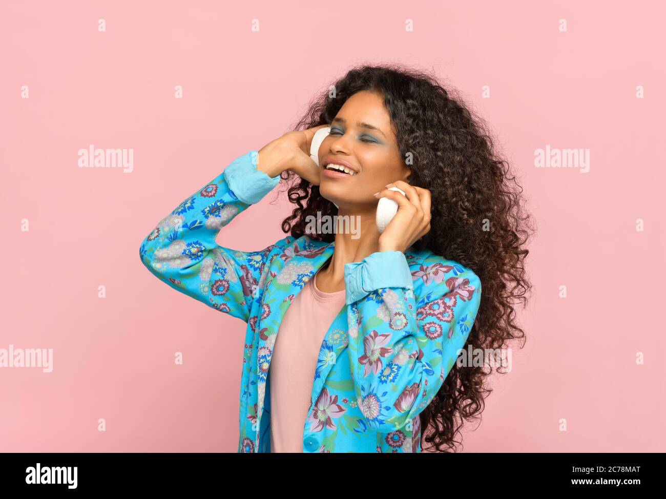 Trendy young Afro-american girl listening to music on stereo headphones with closed eyes and blissful smile against a pink studio background Stock Photo