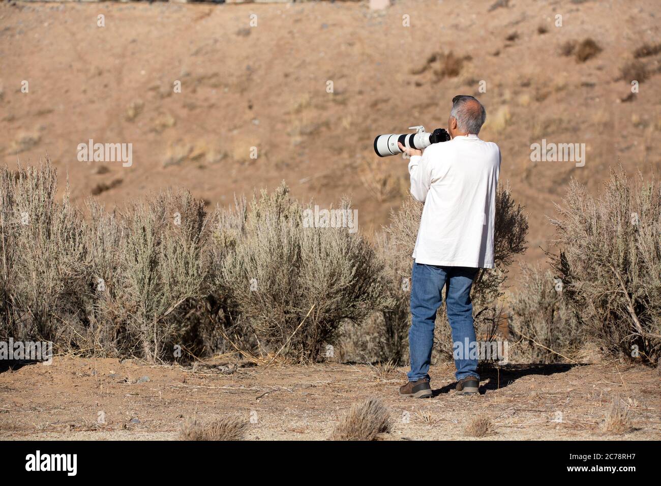 Older man holding a large camera with a white telephoto lens in the desert Stock Photo