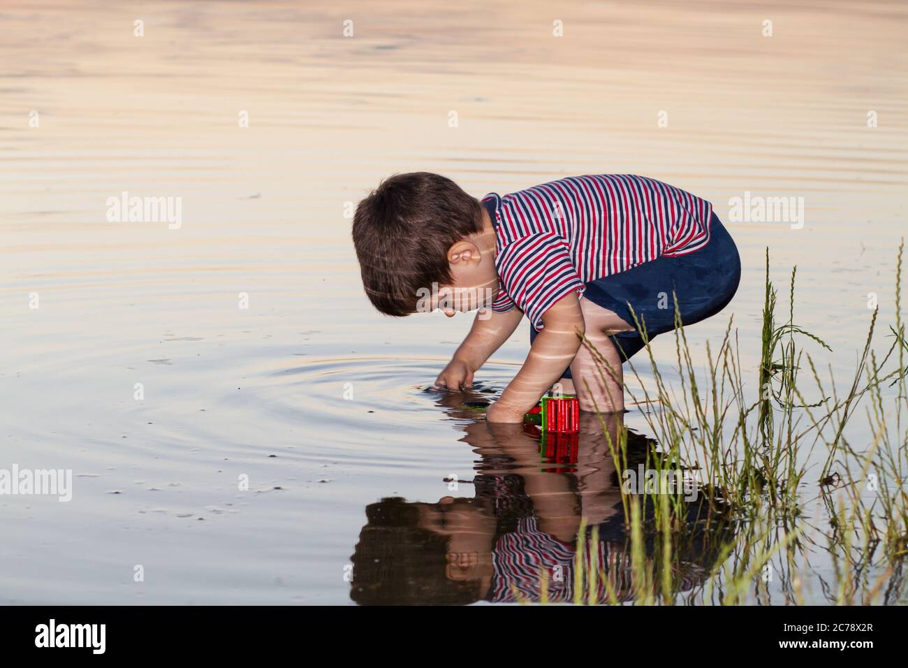 A little boy 2-3 years old plays on the river bank, the reflection of a child in the water. Outdoors in summer. Stock Photo