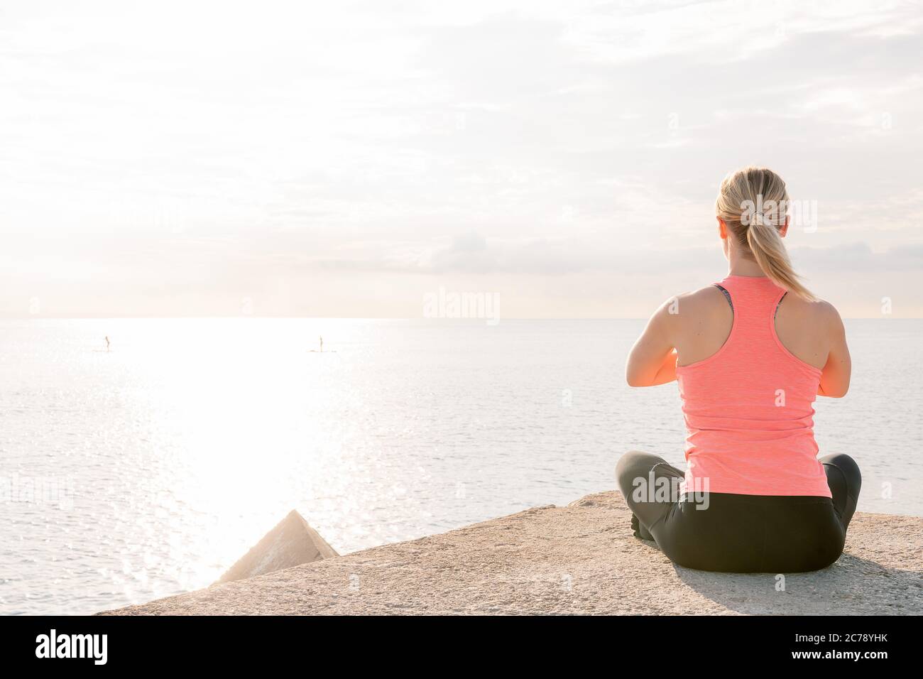 back view of an unrecognizable blonde woman doing yoga at dawn sitting in front of the sea, mental health care and relaxation concept, copy space for Stock Photo