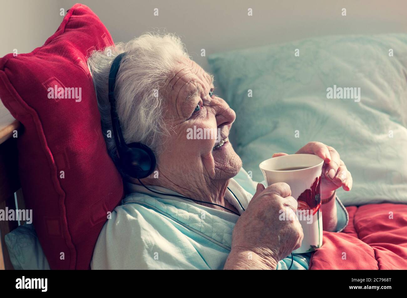 Care home elderly 95 years lady happy resting in bed wearing headphones enjoys a cup of tea in her sunny comfortable care room home Stock Photo