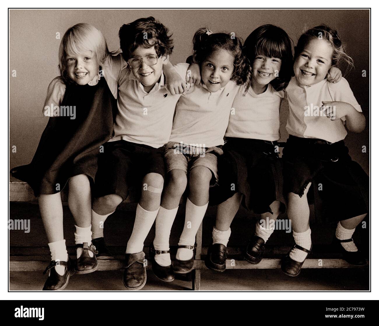 JUNIOR SCHOOL PUPILS RETRO HAPPY DAYS VINTAGE NATURAL INFANTS GROUP 5-7 years  3 girls and 1 boy smiling sitting on school bench,  B&W Kodak Tri-X film study of primary school pupils sitting together on bench in natural light, enjoying being together as friends and having an informal school photo taken.. Stock Photo
