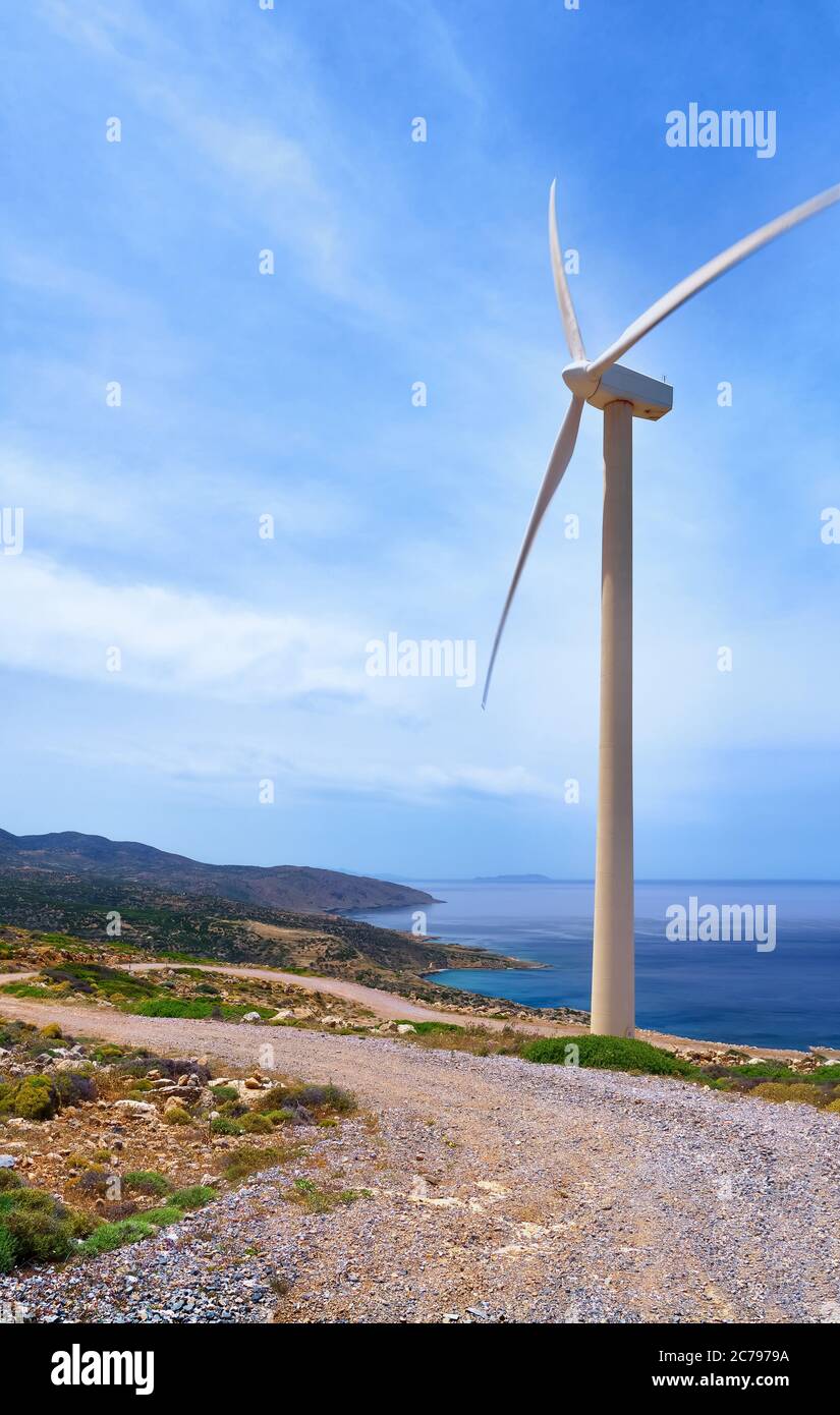 Single windmill turbine on hilltop of seashore in colorful landscape against dynamic blue sky with clouds and winding road. Stock Photo