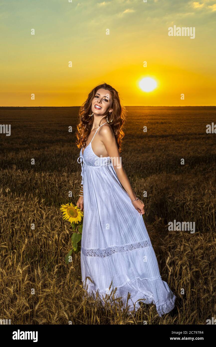 Beautiful woman in white dress in a wheat field with a sunflower flower ...