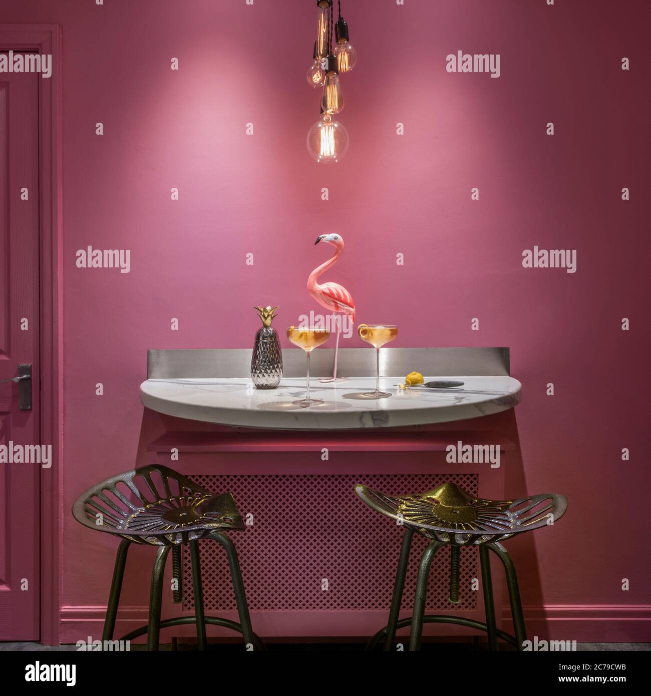 Marble semi-circular breakfast bar in kitchen with pink wall and two cast iron stools. Two cocktail glasses with pineapple shaped cocktail shaker and Stock Photo
