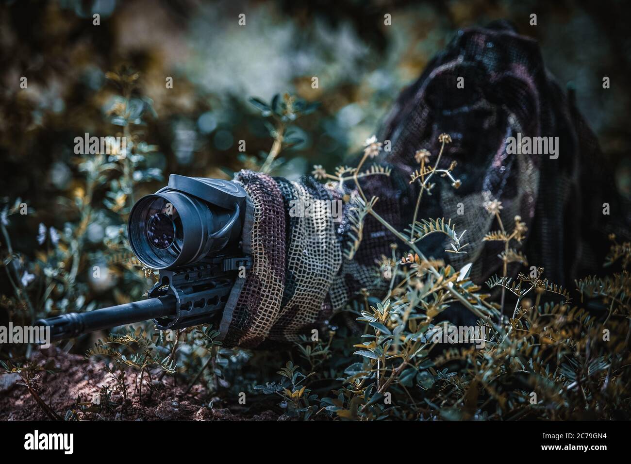 Sniper in camouflage suit in an ambush among the bushes. The shooter aims from a carbine with an optical sight. Sniper in position. Stock Photo