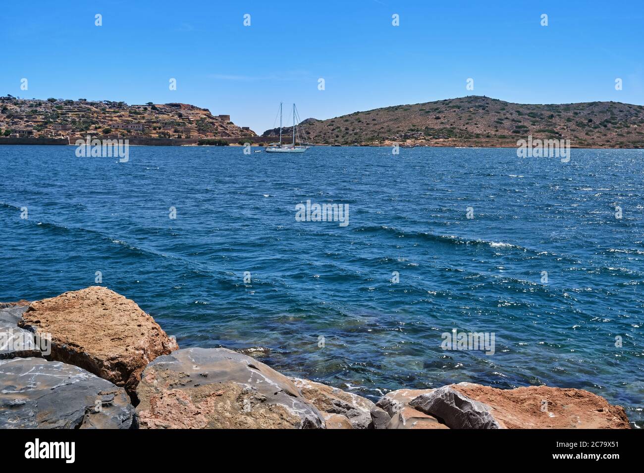 View of Spinalonga island lagoon and Venetian fortress with anchored yacht and breakwater stones in foreground, Crete, Greece Stock Photo