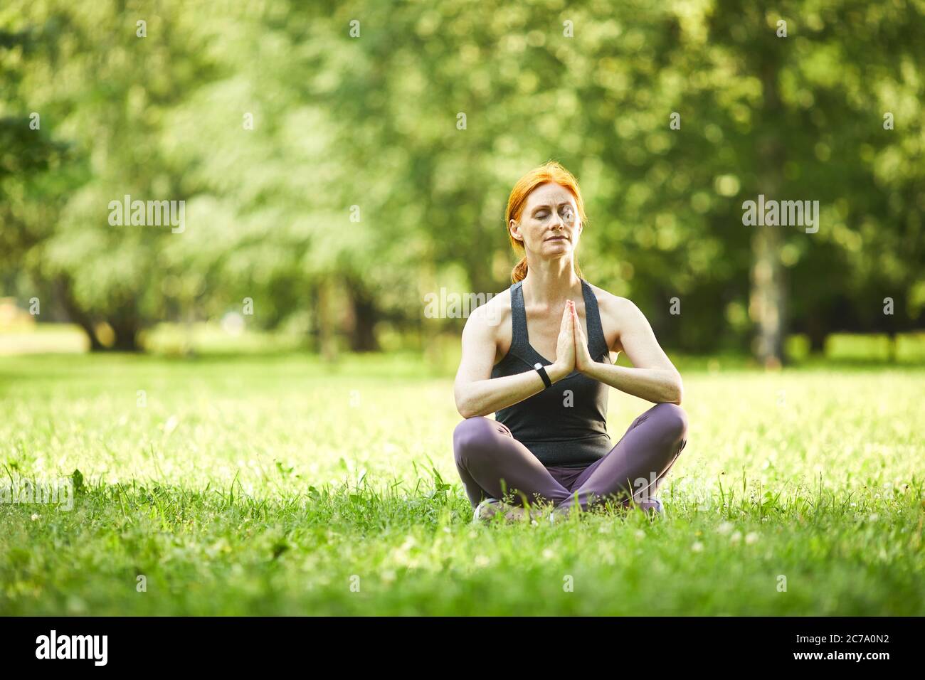 Serene redhead mature woman sitting on grass and holding hands in Namaste while meditating in empty park Stock Photo