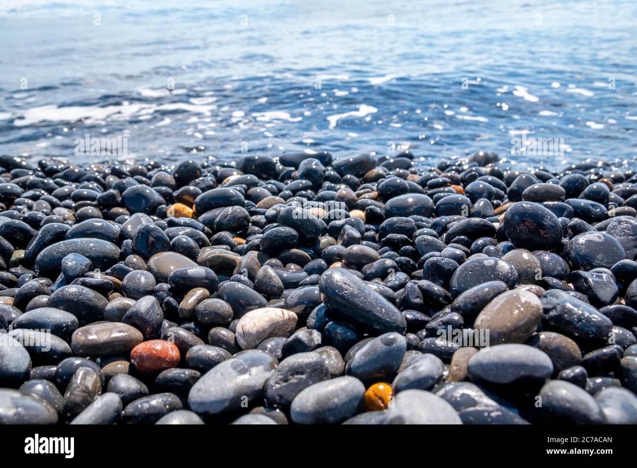 An empty black beach with pebbles and blue turquoise clear, calm water