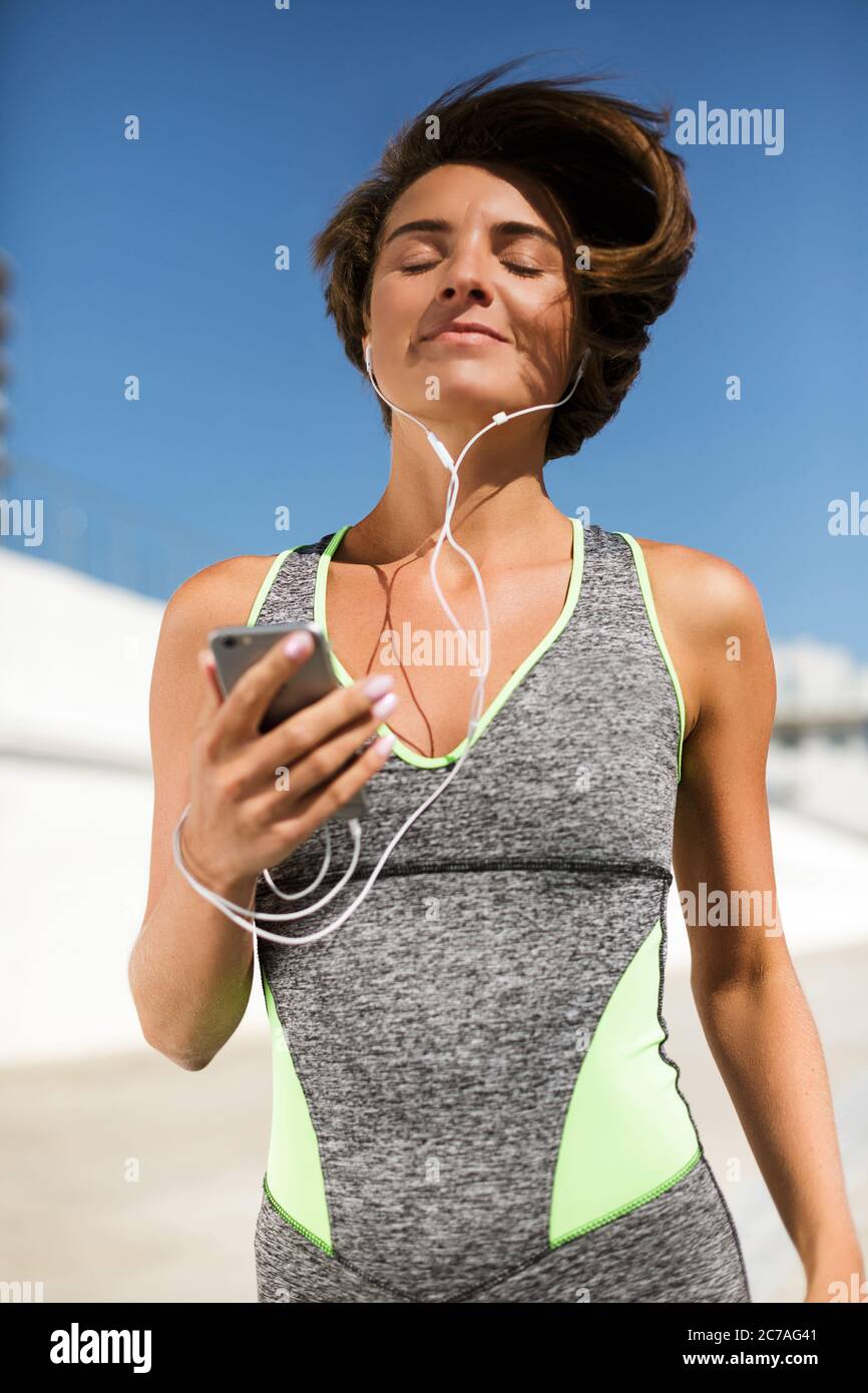 Beautiful smiling woman in modern gray sport suit jogging holding cellphone and earphones dreamily Stock Photo