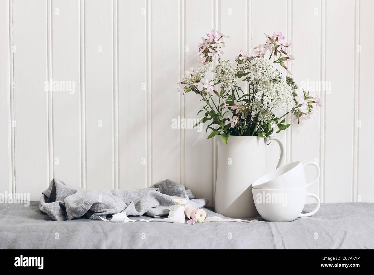 Bouquet of pink soapwort and wild carrot flowers in ceramic pitcher. Empty coffee cups on linen table cloth. White wooden wall background. Feminine Stock Photo