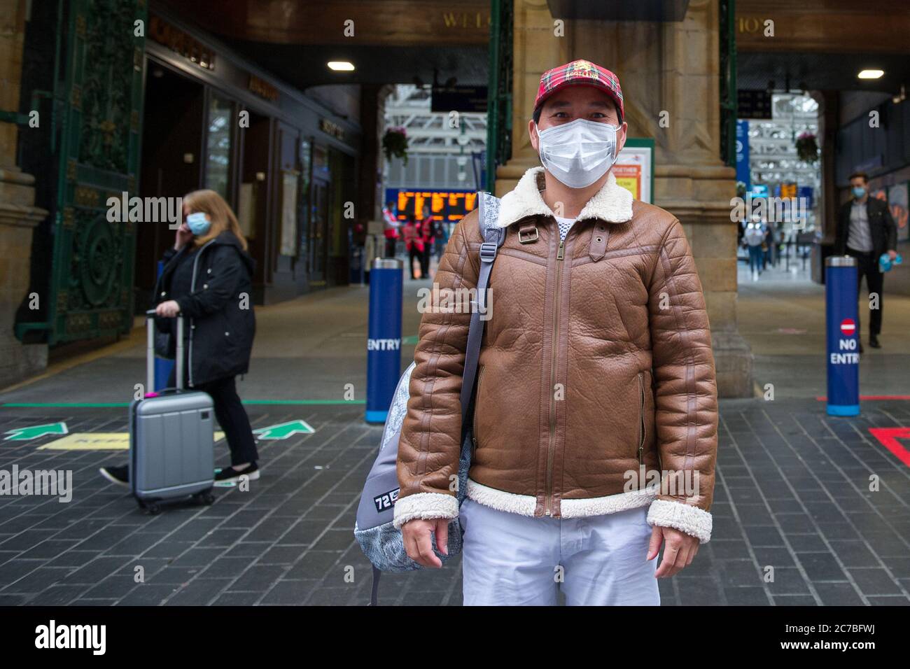Glasgow, Scotland, UK. 16th July, 2020. Pictured: A tourist seen wearing a blue surgical face mask with a royal Stewart tartan baseball cap and brown leather bomber jacket, poses for a picture outside of Glasgow Central station. The Scottish Government made it mandatory that face coverings are worn when travelling on public transport in Scotland to help prevent the spread of coronavirus. Credit: Colin Fisher/Alamy Live News Stock Photo