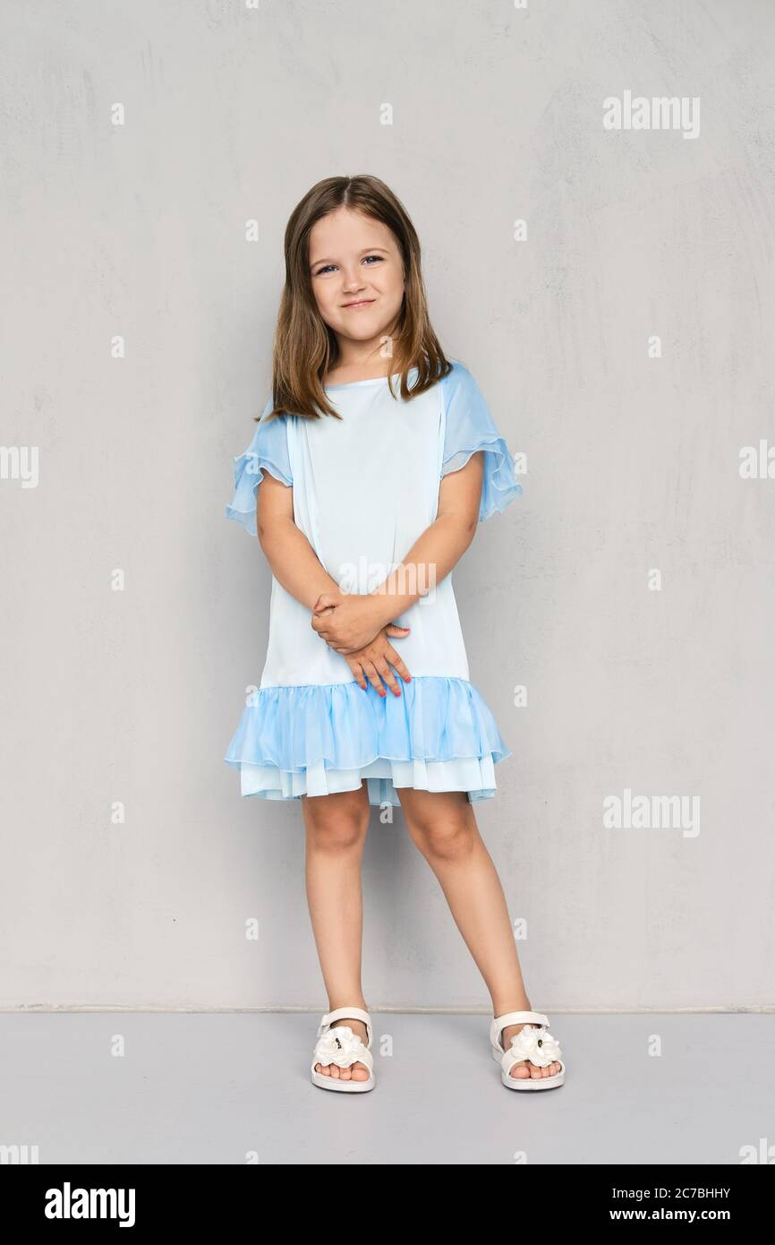 Cute little girl in blue dress and white sandals posing with hands clasped  down near the gray wall Stock Photo - Alamy