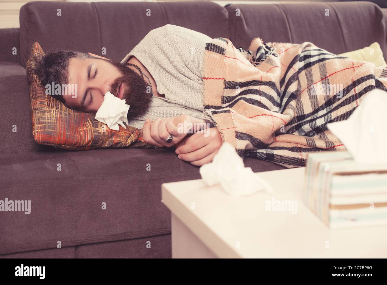 Sick bearded man lying on sofa at home and blowing nose. Stock Photo