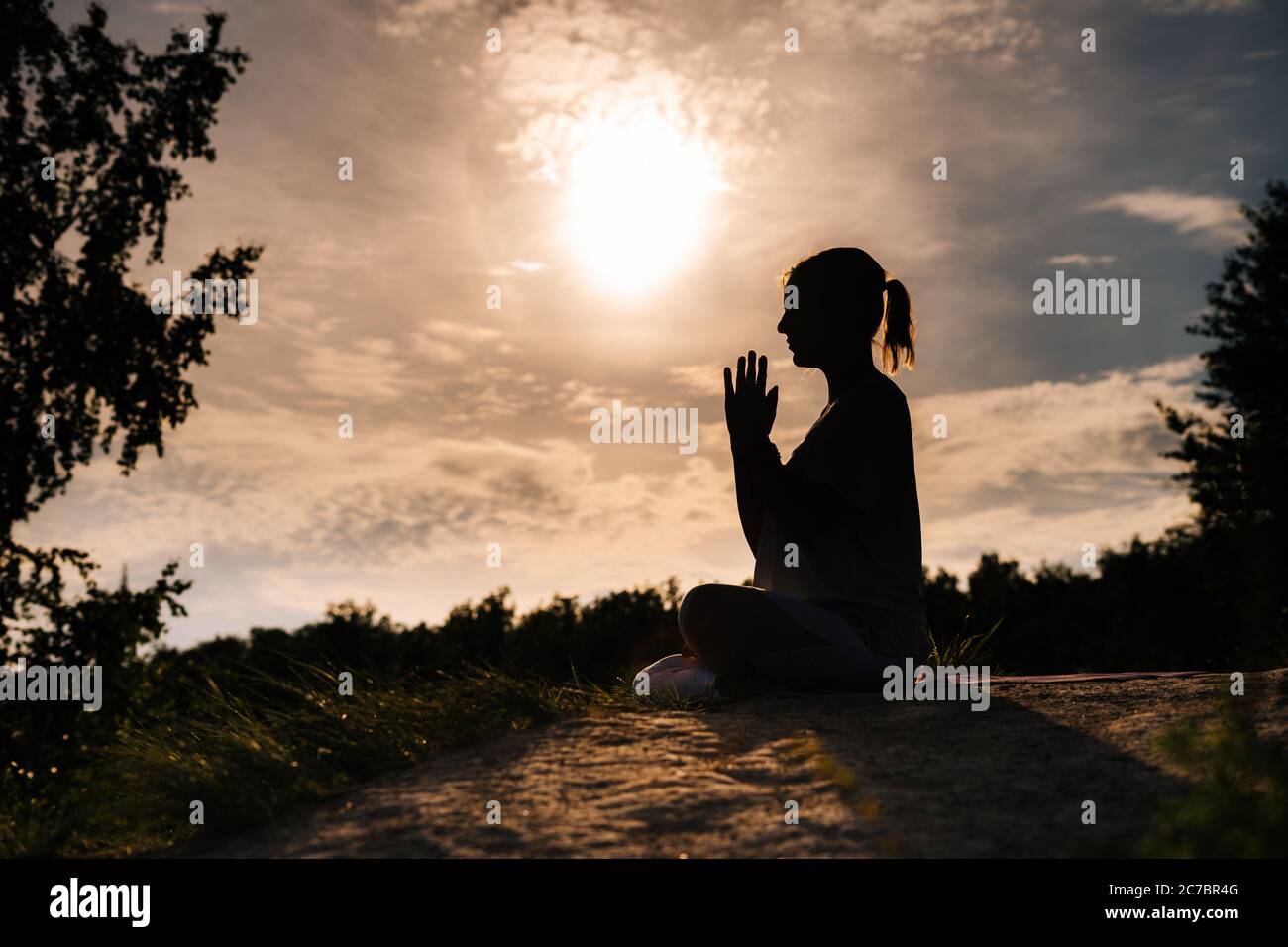 Relaxed lady is performing namaste yoga pose background of sun outdoor on park outside city evening. Stock Photo