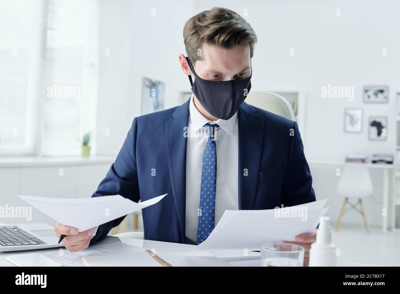 Modern young businessman in cloth mask sitting at table in office and reading contract while examining terms and conditions Stock Photo