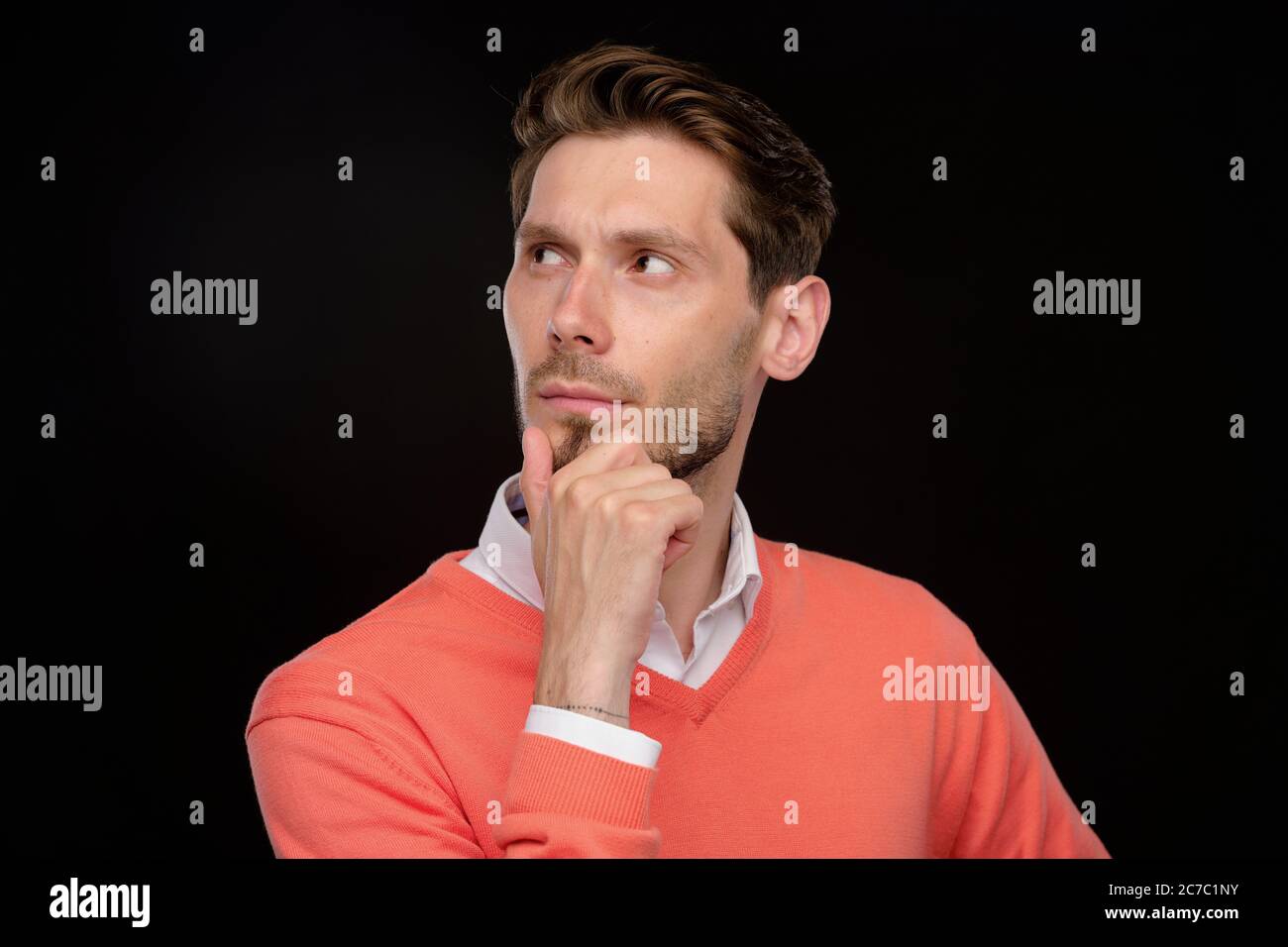 Pensive handsome young man in orange sweater rubbing chin while thinking of strategy, black background Stock Photo