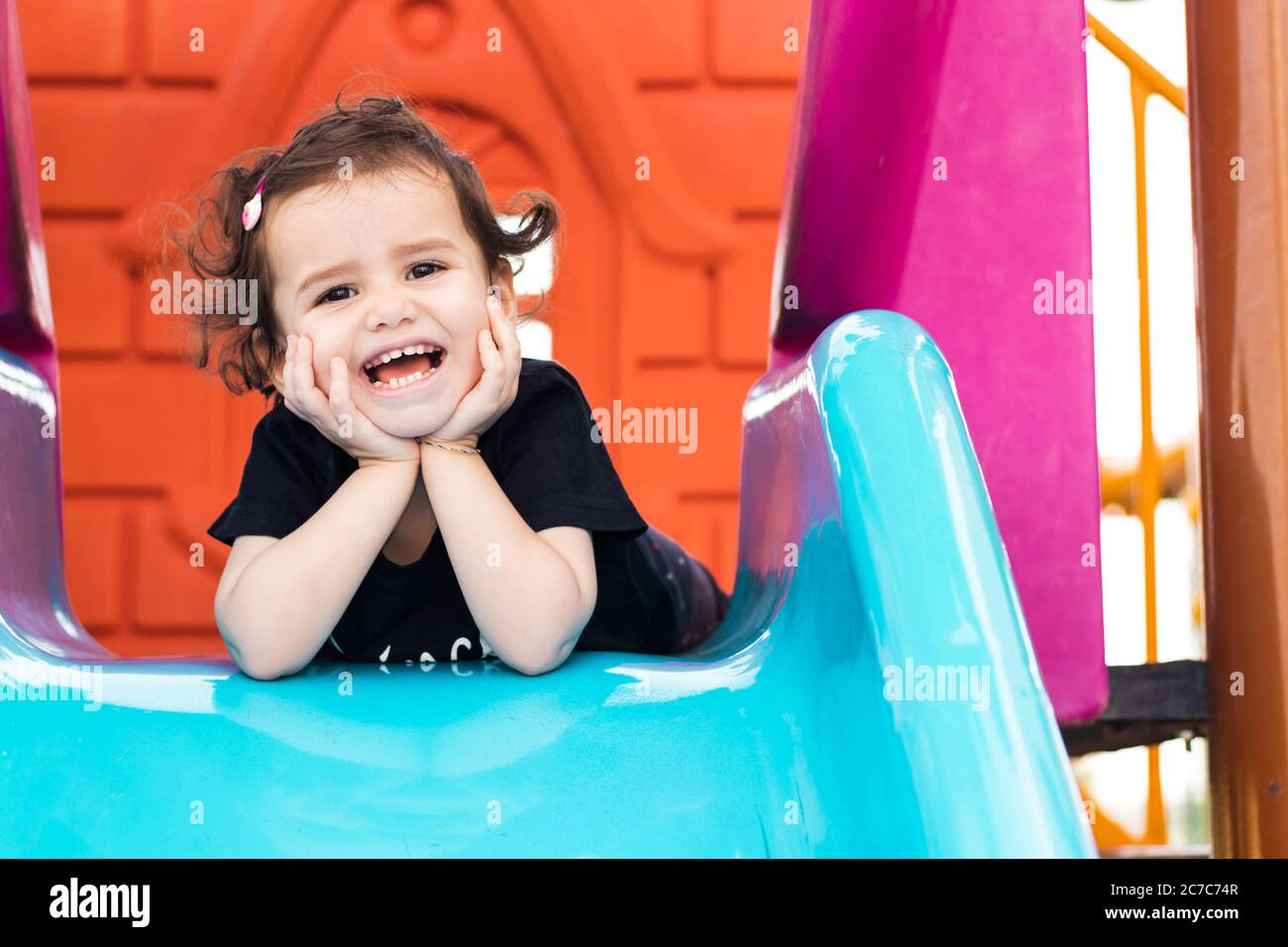 a cute liitle girl laying on the blue slide in a playground with hands under her chin. Stock Photo