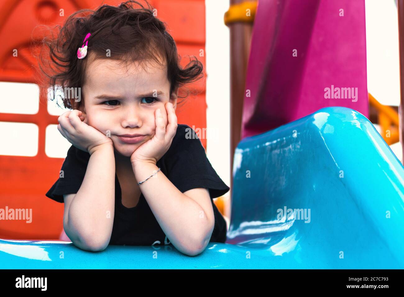 a cute liitle girl laying on the blue slide in a playground with hands under her chin. Stock Photo