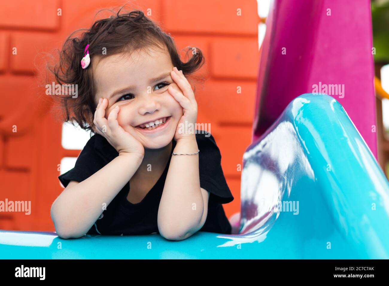 a cute little girl laying on the blue slide in a playground with hands under her chin. Stock Photo