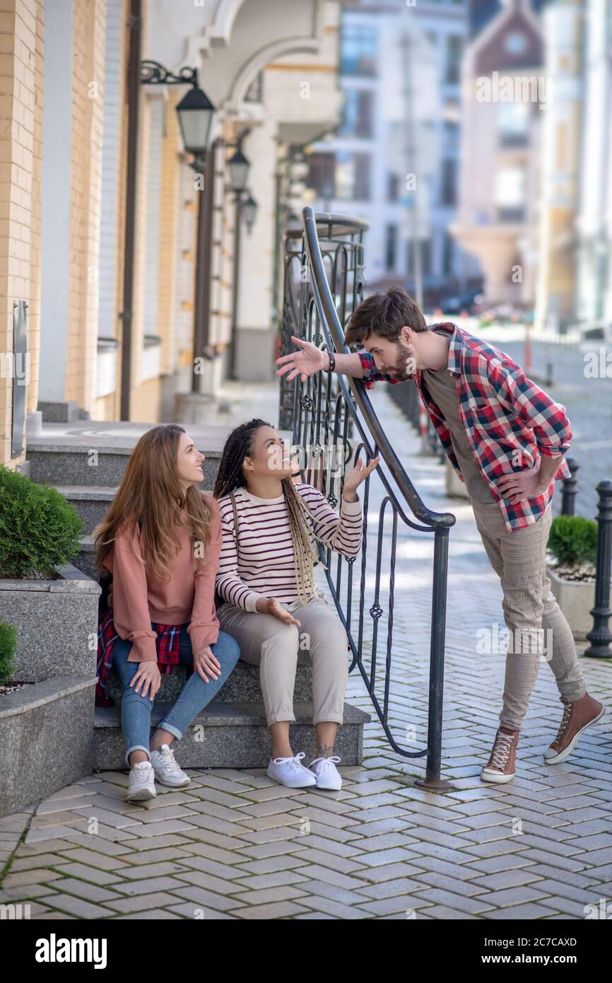 Two girls sitting on steps, guy standing next Stock Photo - Alamy