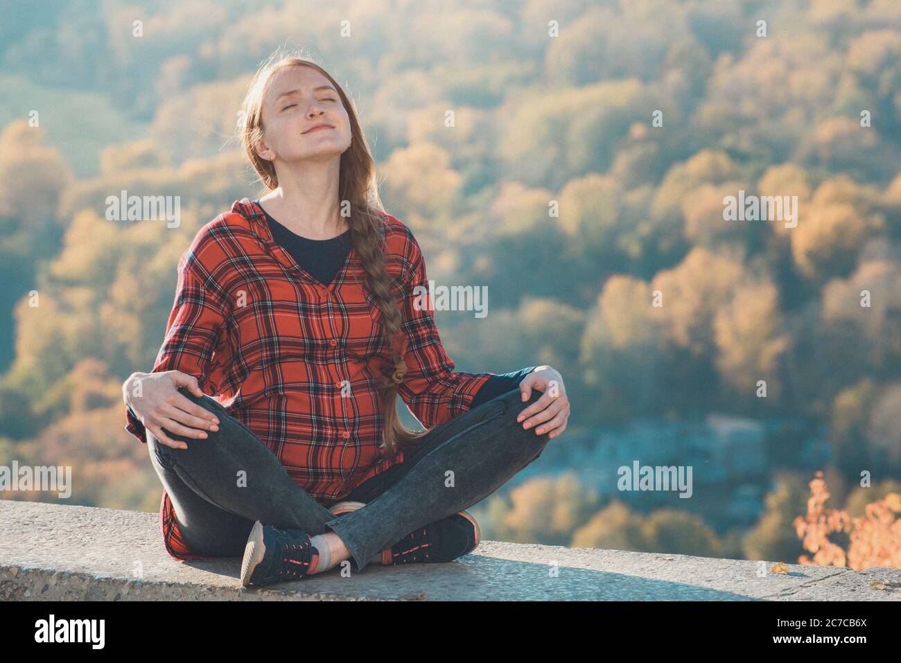 Pregnant woman sits on a hill with her eyes closed. Meditation. Autumn forest on the background Stock Photo