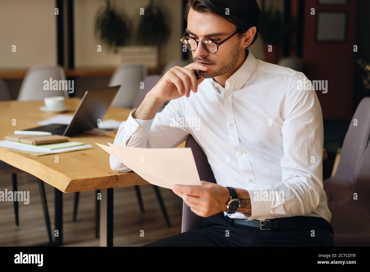 Young serious businessman in eyeglasses thoughtfully reading papers while working in modern office Stock Photo