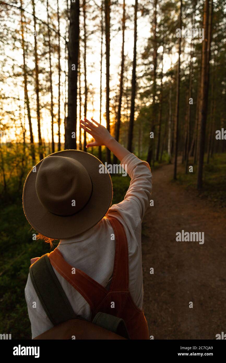Woman naturalist in hat and orange overalls with backpack looking at the sun through her fingers at sunset, exploring wildlife and ecotourism adventur Stock Photo