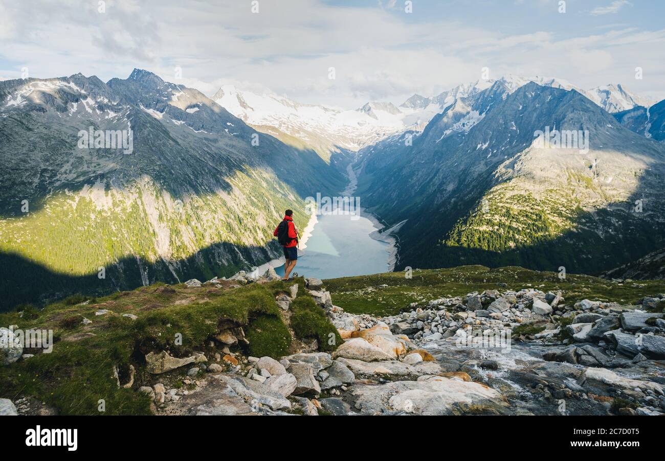 Lonely man with backpack walking on bridge the top of the mountain and looking at beautiful landscape. In the background a glacial dam during sunset Stock Photo