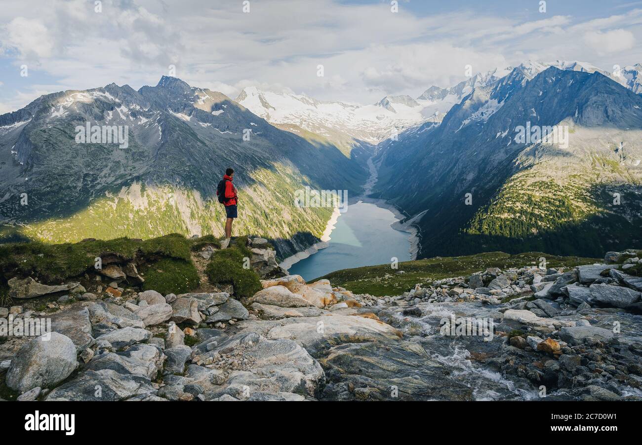 Adventurous man on the edge of a cliff overlooking the beautiful Austria Alps and Lake during a vibrant summer sunset. Taken in Hiking to the Olperer Stock Photo
